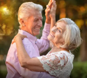 A happy older couple with white hair, dancing and smiling at each other in an outdoor setting with blurred trees and sunlight.